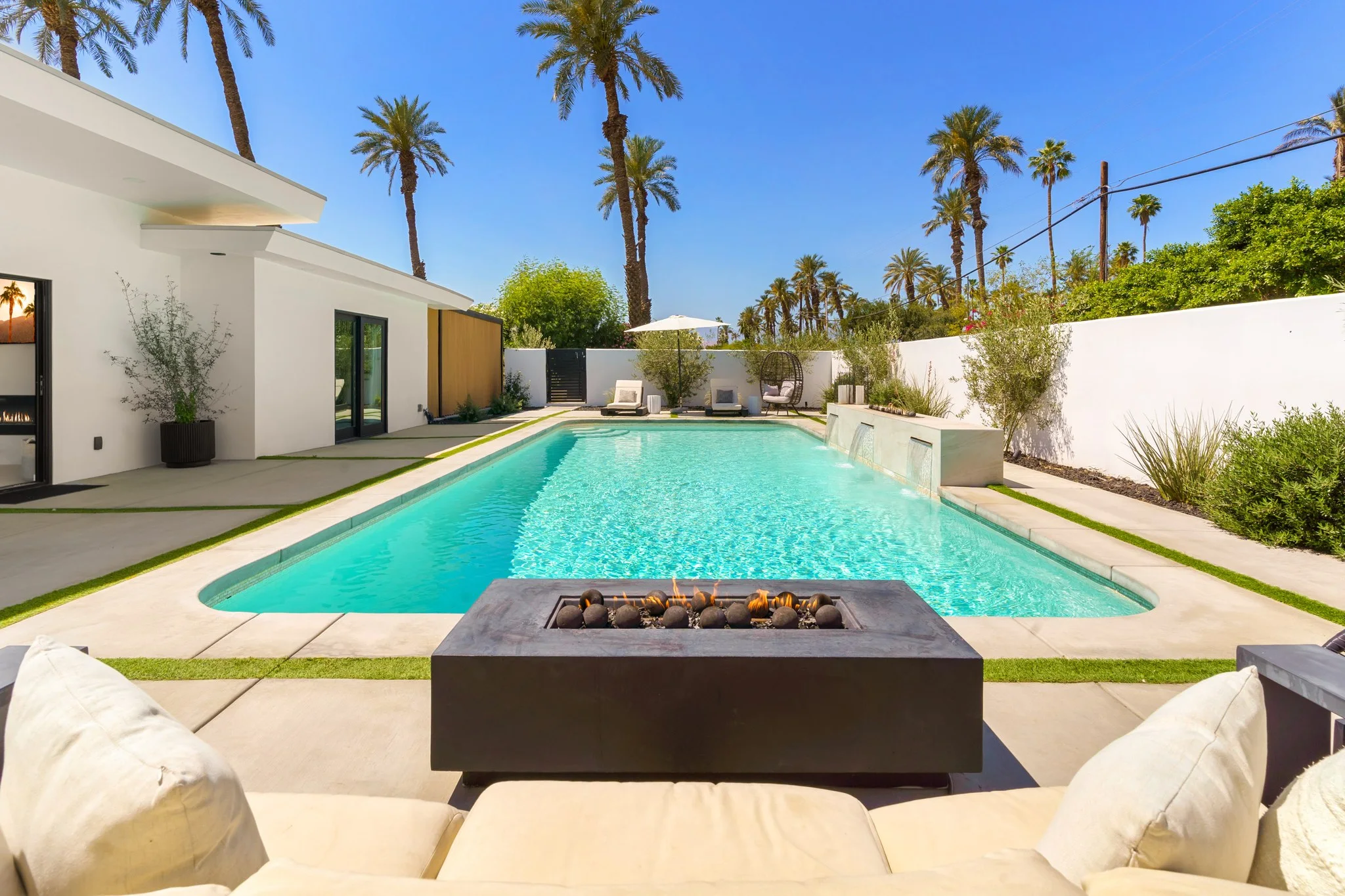 Long pool and black fire table under palms at midday, white garden walls around the yard