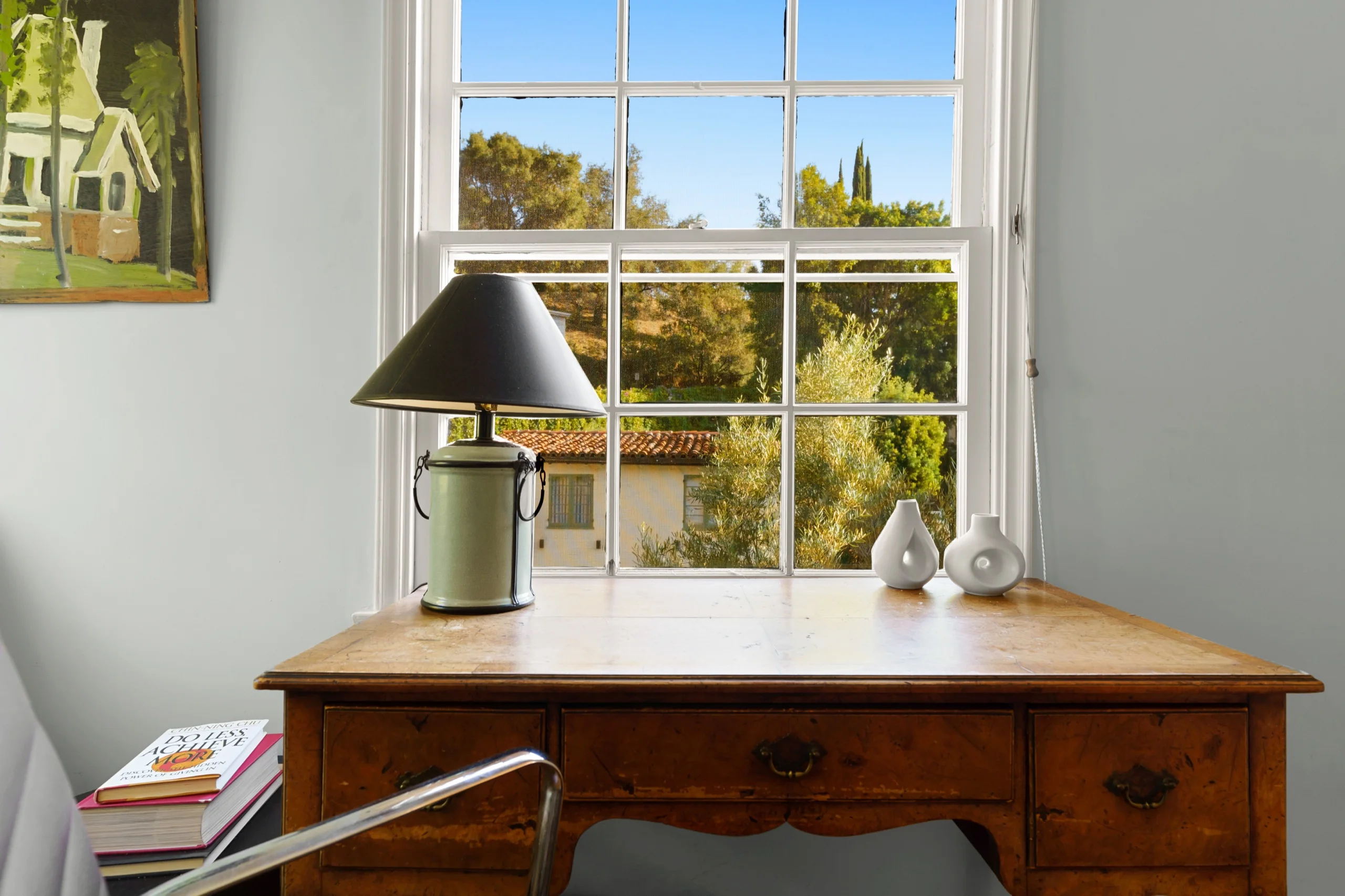 Desk detail — a brass lamp and a pair of small vases at a sash window looking into the trees