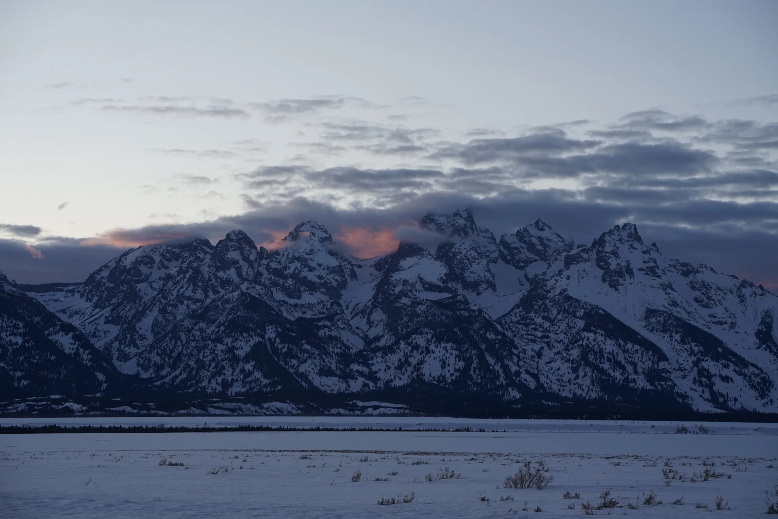 Snow-covered Teton range under heavy cloud at dusk, flat white valley in the foreground